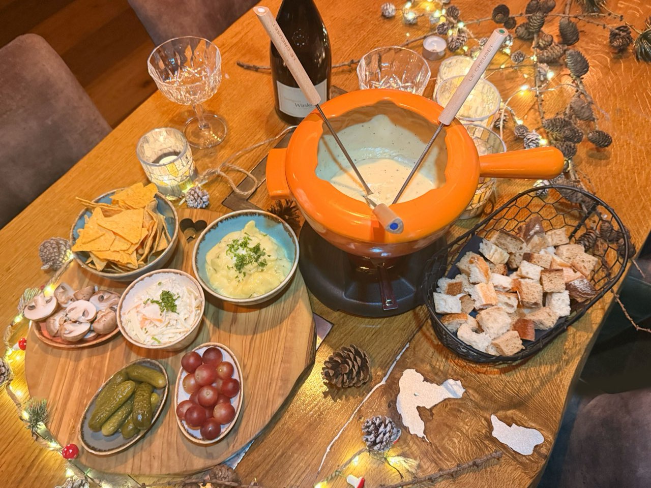 Käsefondue im orangefarbenen Topf mit Brotwürfeln, Trauben, Gurken und Dips