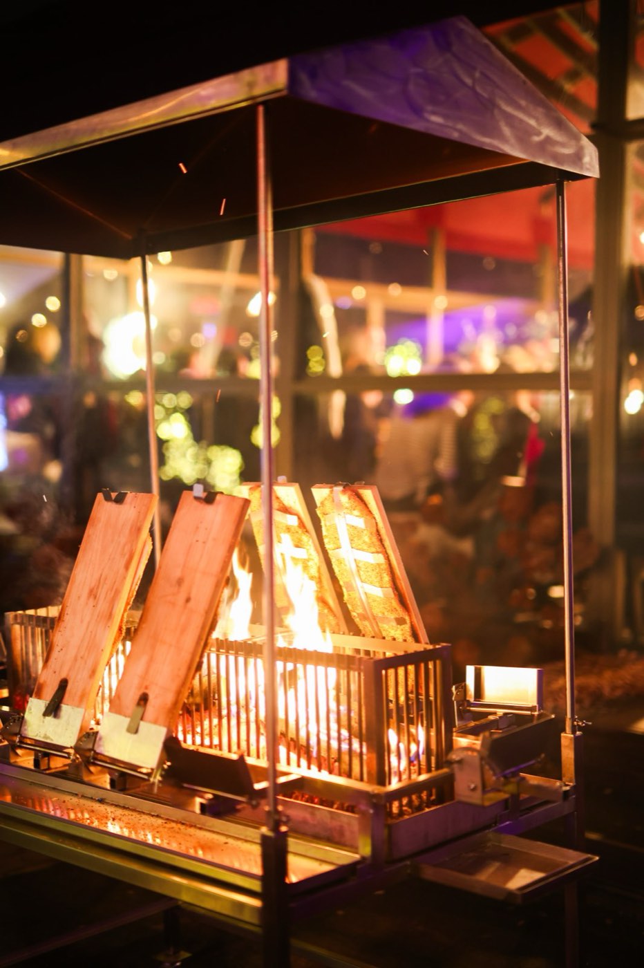 Flammlachs-Grill bei Nacht: Lachsseiten auf Holzbrettern stehen schräg um ein offenes Feuer, darüber ein kleines Dach, im Hintergrund das beleuchtete Winterzelt