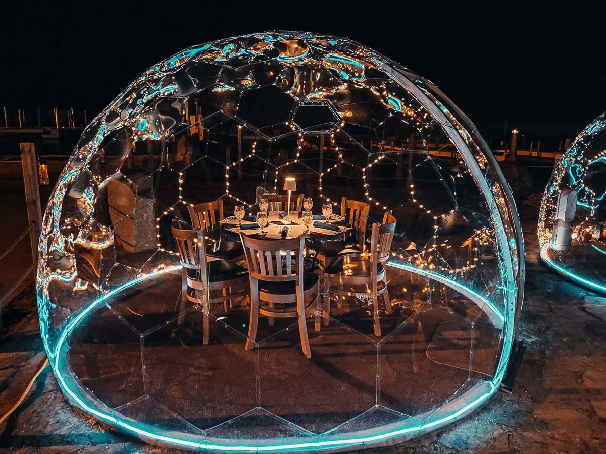 Klare Kuppel bei Nacht mit Leuchtband am Boden, innen ein gedeckter Tisch mit Stühlen und Lichterketten
