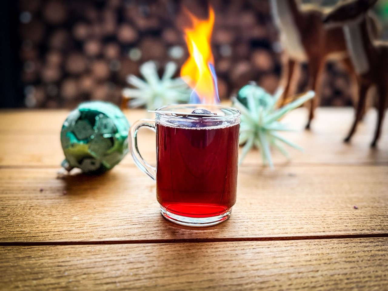 Glas mit rotem Heißgetränk, obenauf brennt eine Flamme, daneben Weihnachtskugeln
