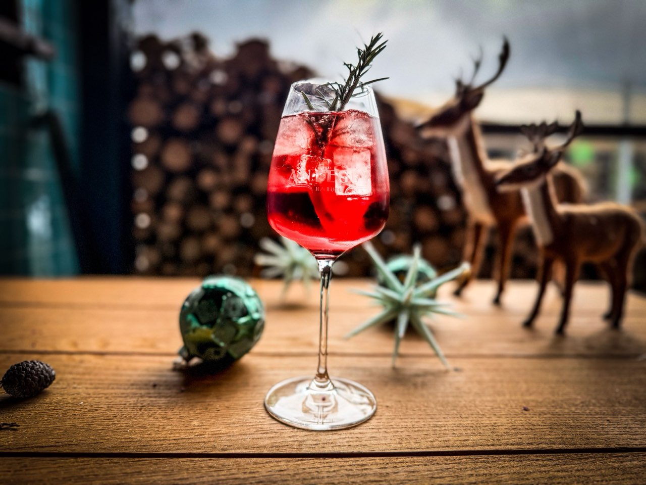 Roter Cocktail mit Eis und Rosmarinzweig im Weinglas, dahinter kleine Hirschfiguren