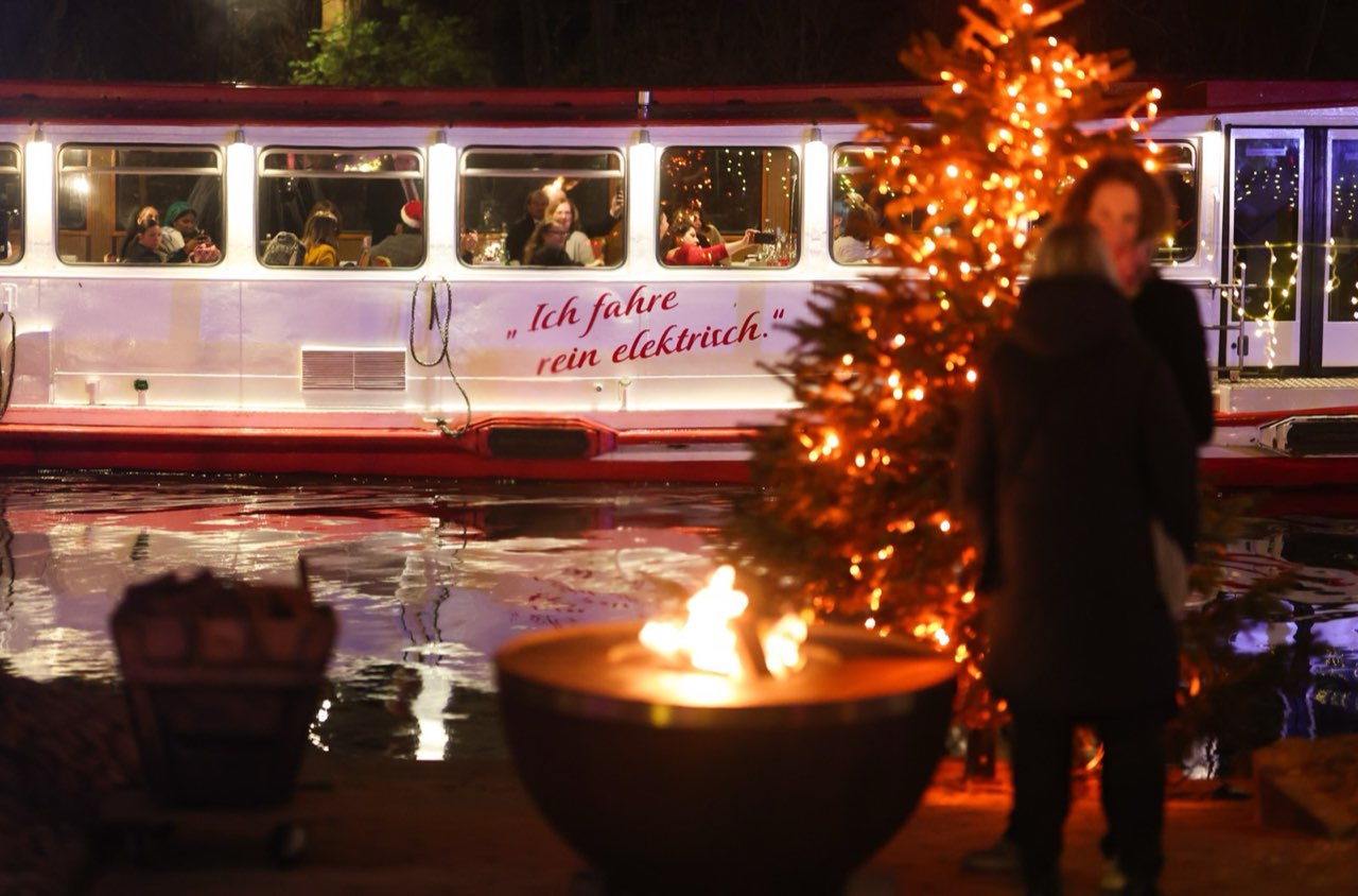 Geschmückter Tannenbaum und Feuerschale am Ufer, dahinter fährt ein beleuchteter Alsterdampfer vorbei