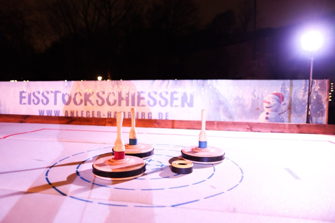 Eisstöcke auf der beleuchteten Bahn, dahinter die Bande mit der Aufschrift Eisstockschiessen