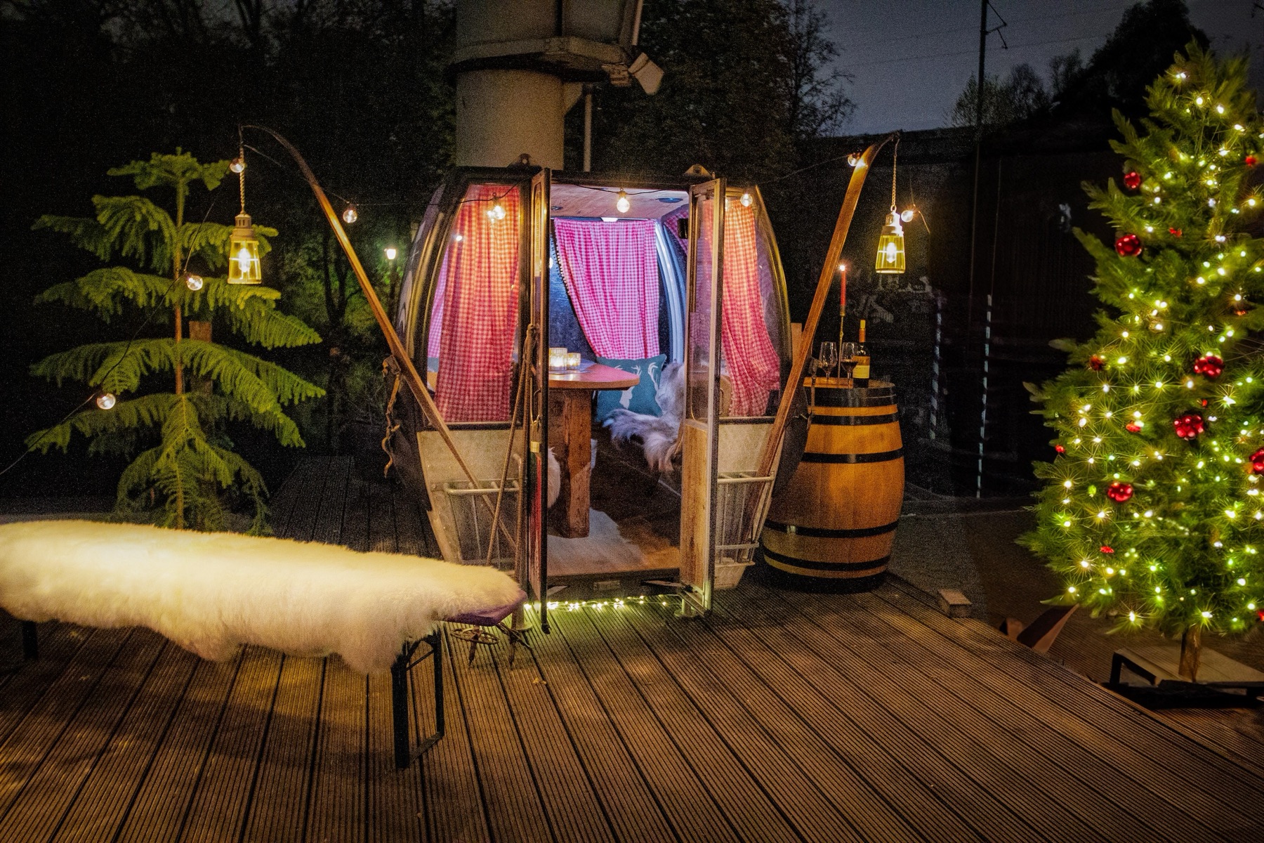 Ski-Gondel bei Nacht auf dem Holzdeck am Wasser, Tür offen, innen ein gedeckter Tisch mit rot karierten Vorhängen, daneben ein geschmückter Tannenbaum